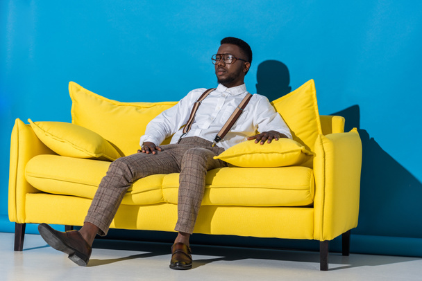 handsome stylish young african american man sitting on yellow couch and looking away on blue   - Foto, Imagem