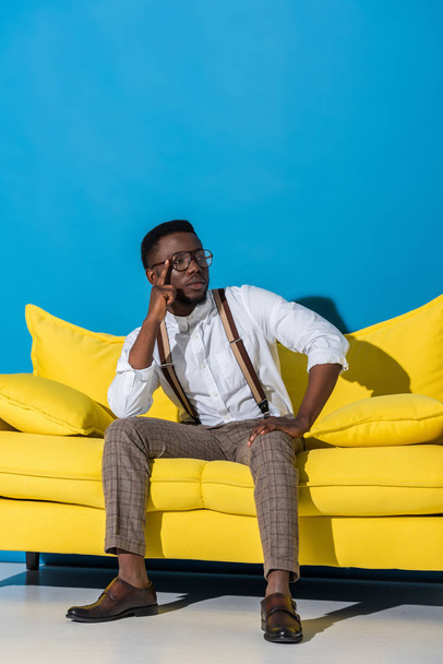 handsome stylish young african american man sitting on yellow couch on blue  - Fotoğraf, Görsel