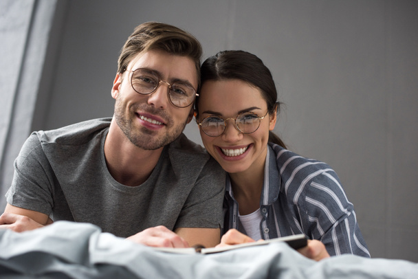 Uśmiechający się para w okularach pisania w Notatniku podczas leżenia w łóżku - Zdjęcie, obraz