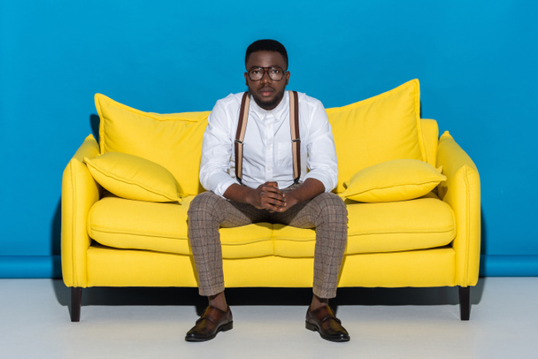 joven afroamericano hombre en gafas sentado en sofá y mirando a la cámara
  - Foto, imagen