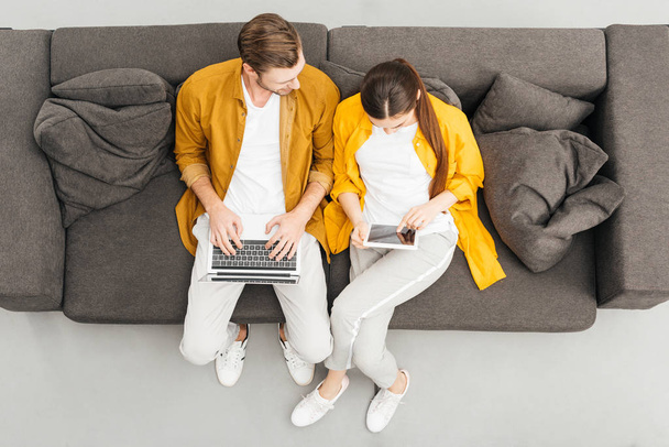 Genç çiftin evde rahat kanepede dijital cihazlar kullanarak havai görünümü - Fotoğraf, Görsel
