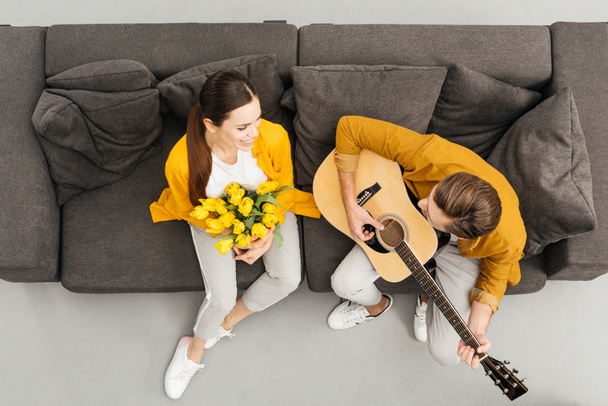 yleisnäkymä mies soittaa kitaraa tyttöystävänsä kun hän tilalla kimppu sohvalla kotona
 - Valokuva, kuva