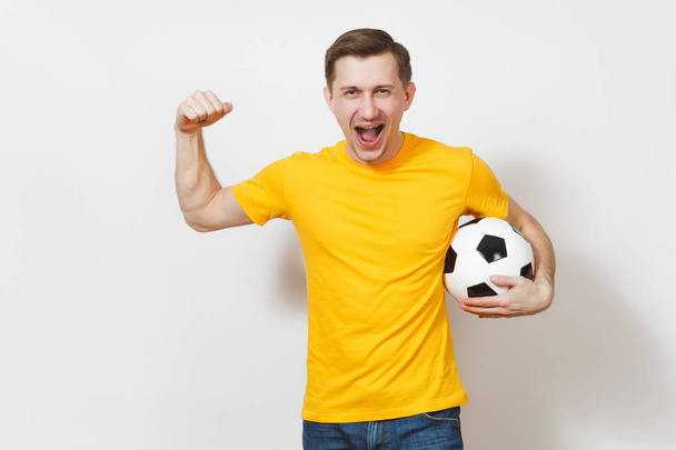 Geïnspireerd Europese jongeman, ventilator of speler in geel uniform houdt van voetbal, juichen favoriete voetbalteam, expressieve zwaaien handen geïsoleerd op een witte achtergrond. Lifestyle concept. Kopiëren van ruimte. - Foto, afbeelding