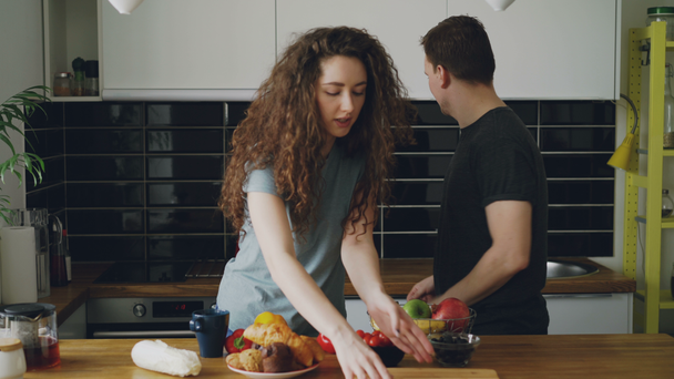 young caucasian loving couple kissing and putting different vegetables and fruits on table to cut them, man giving knife to woman, they are nice and positive - Πλάνα, βίντεο