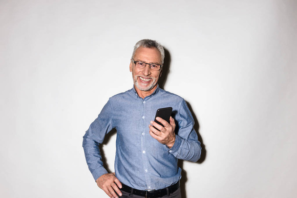 Cheerful mature man smiling and holding smartphone isolated - Foto, imagen