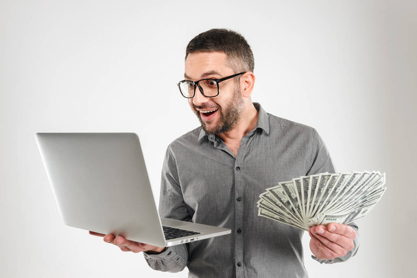 Excited businessman using laptop computer holding money. - Zdjęcie, obraz