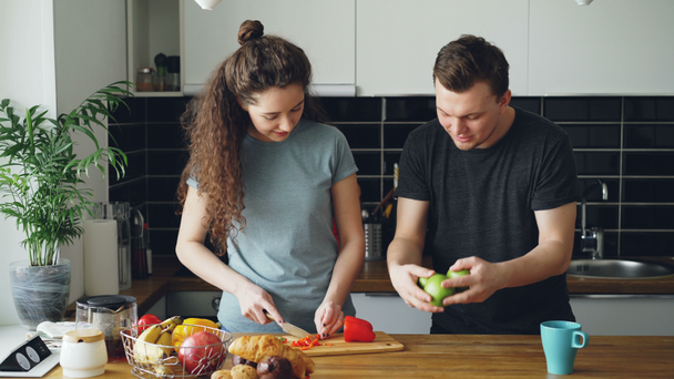 Cheerful woman preparing breakfast in the kitchen, her husband comes taking apples and showing her juggling, woman laughing, one apple falls and couple looking for apple - Imágenes, Vídeo