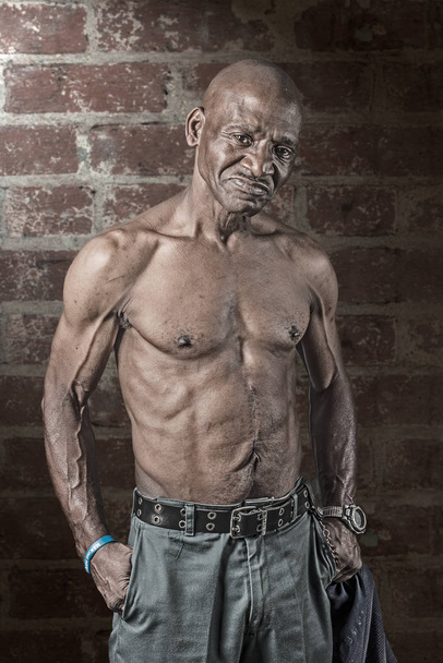 Kova lihaksikas vanhempi afroamerikkalainen mies ilman paitaa dramaattisesti valaistu tiiliseinää vasten
 - Valokuva, kuva