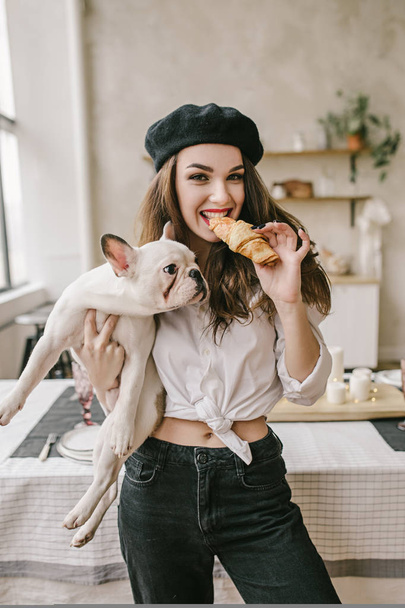 Portrait of a happy woman wearing black beret holding croissant and french puppy bulldog - Φωτογραφία, εικόνα
