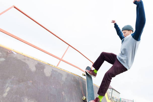 Teen skater in a hoodie sweatshirt and jeans slides over a railing on a skateboard in a skate park - 写真・画像