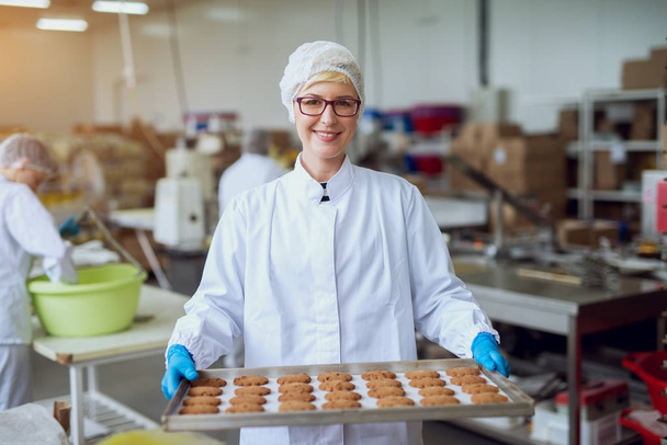 joven feliz trabajadora sosteniendo galletas recién horneadas en hojalata en la fábrica de producción de alimentos
 - Foto, Imagen