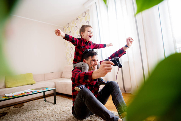 веселий збуджений батько і маленький син в червоних сорочках грають в консольну гру з геймпадами
 - Фото, зображення