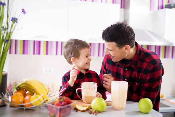 Junge fröhliche Vater und Sohn in roten Hemden kochen gesundes Frühstück mit Mixer  - Foto, Bild