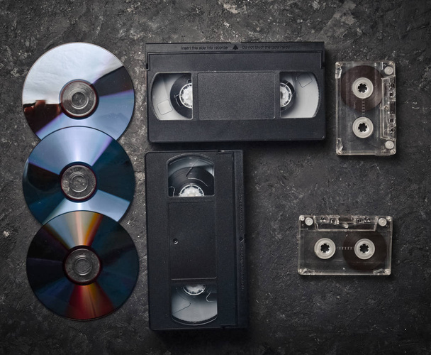 Cámaras de vídeo planas, CD, cassette de audio sobre una superficie de hormigón negro. Tecnología de medios retro del pasado. Vista superior
. - Foto, Imagen