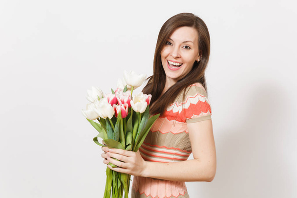 Beautiful young happy brunette female in light patterned dress is holding bouquet of white and pink tulips in hands, smiling and rejoices isolated on white background. Concept of holiday, good mood. - Photo, Image