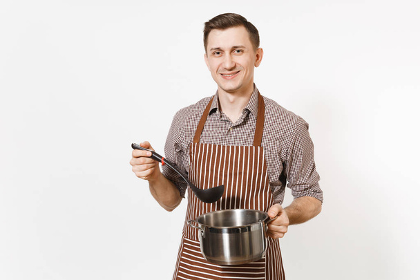 Man chef in striped brown apron holding silver stainless glossy aluminium empty stewpan, pan or pot, ladle isolated on white background. Male housekeeper or houseworker. Kitchenware, dishes concept. - Photo, Image