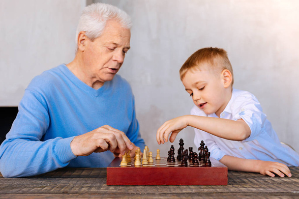 Pleasant aged man playing chess with his grandson - Fotó, kép