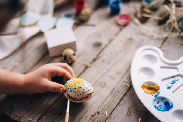 Oříznutý pohled dítěte ruce drží a barvení vajec v tečky s kartáčem a modré a žluté barvy na dřevěný stůl s nástroji pro umění - Fotografie, Obrázek