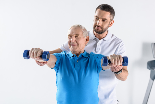 портрет реабилитационного терапевта, помогающего старшему мужчине тренироваться с гантелями на сером фоне
 - Фото, изображение