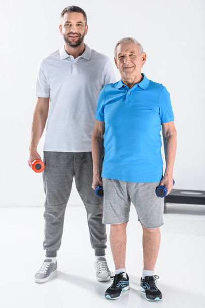 terapeuta de rehabilitación y hombre mayor con mancuernas mirando a la cámara sobre fondo gris
 - Foto, Imagen