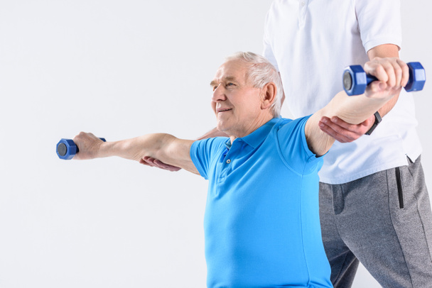 Teilansicht eines Reha-Therapeuten, der einem älteren Mann beim Training mit Kurzhanteln auf grauem Hintergrund hilft - Foto, Bild