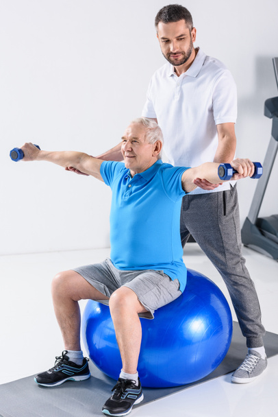 rehabilitation therapist assisting senior man exercising with dumbbells on fitness ball - Photo, Image