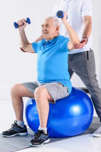 przycięte strzał rehabilitacji terapeuta pomoc starszy człowiek, ćwiczenia z hantlami na piłka fitness na szarym tle - Zdjęcie, obraz