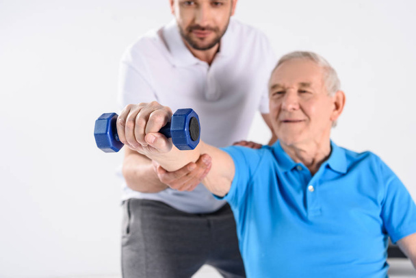 selective focus of rehabilitation therapist assisting senior man exercising with dumbbell isolated on grey - Photo, Image