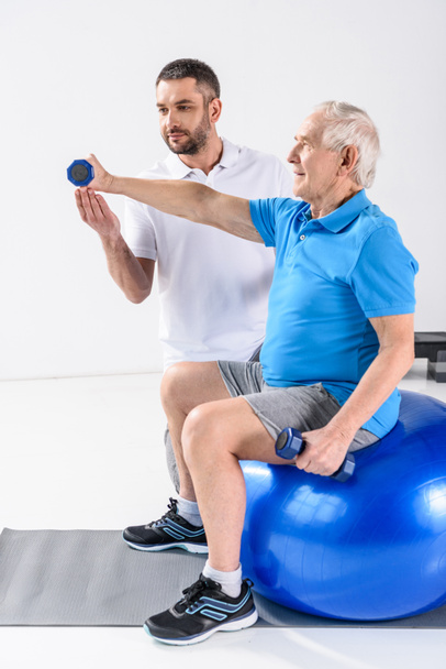 rehabilitation therapist assisting senior man exercising with dumbbells on fitness ball - Фото, зображення