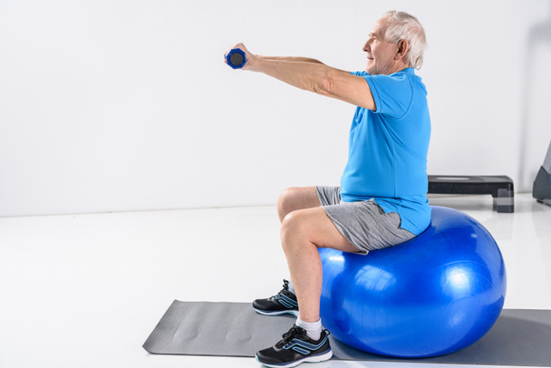 side view of senior man with dumbbells exercising on fitness ball on grey backdrop - Photo, Image