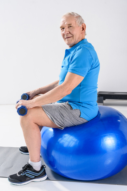 side view of senior man with dumbbells sitting on fitness ball on grey backdrop - 写真・画像