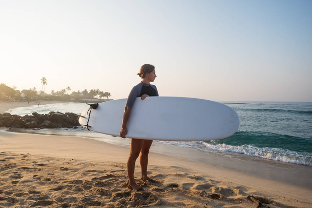 mujer caucásica con tabla de surf de pie cerca del agua y mirando las olas
. - Foto, imagen