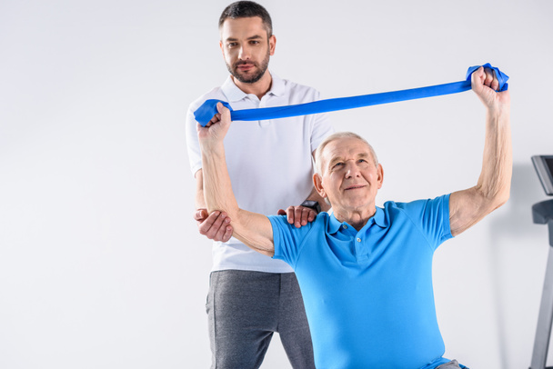 portrait of rehabilitation therapist assisting senior man exercising with rubber tape on grey backdrop - 写真・画像