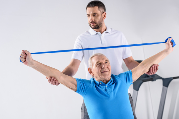 retrato de terapeuta de reabilitação auxiliando o homem idoso a se exercitar com fita de borracha
 - Foto, Imagem
