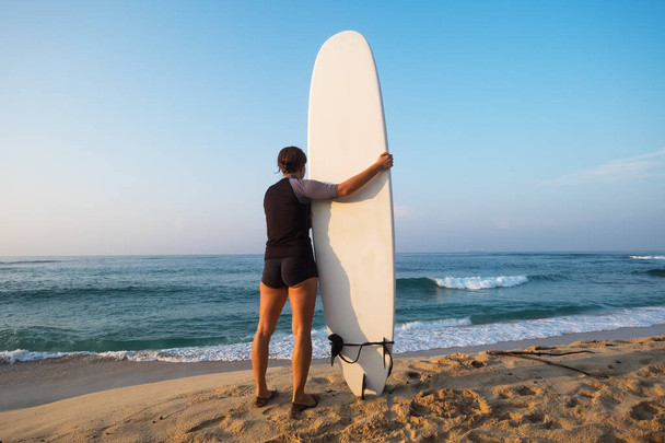 біла жінка з дошкою для серфінгу, що стоїть біля води і дивиться на хвилі
. - Фото, зображення