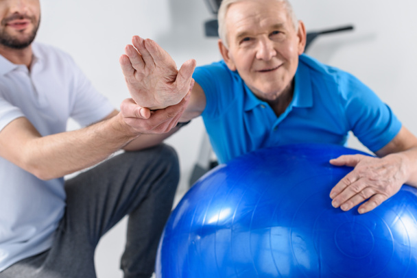 vue partielle du thérapeute de réadaptation aidant le vieil homme souriant faisant de l'exercice sur le ballon de fitness
 - Photo, image