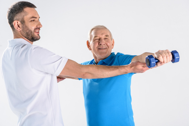 Porträt eines Rehabilitationstherapeuten, der einem älteren Mann beim Training mit einer Hantel hilft, isoliert auf grau - Foto, Bild