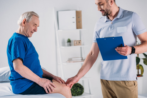 side view of rehabilitation therapist with notepad checking senior mans knee on massage table - Foto, imagen