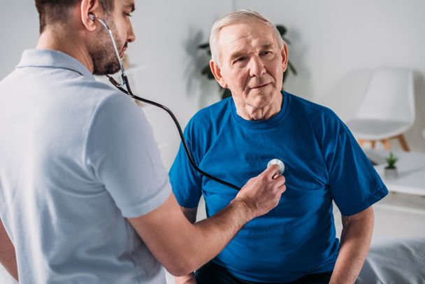 rehabilitation therapist with stethoscope checking senior mans heartbeat - Foto, afbeelding