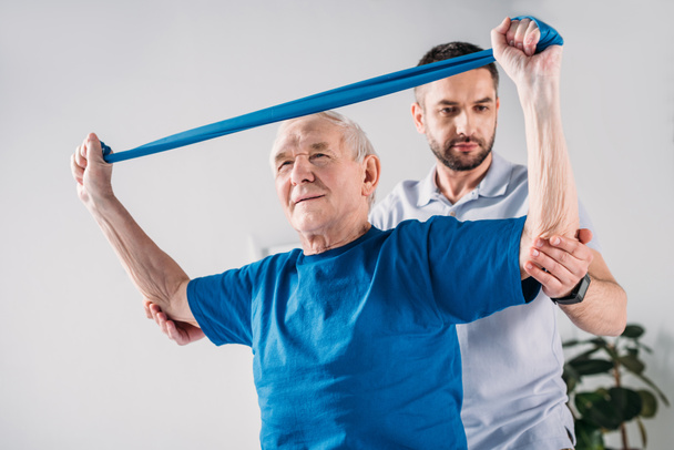 terapeuta de reabilitação focado ajudando o homem idoso a se exercitar com fita de borracha
 - Foto, Imagem