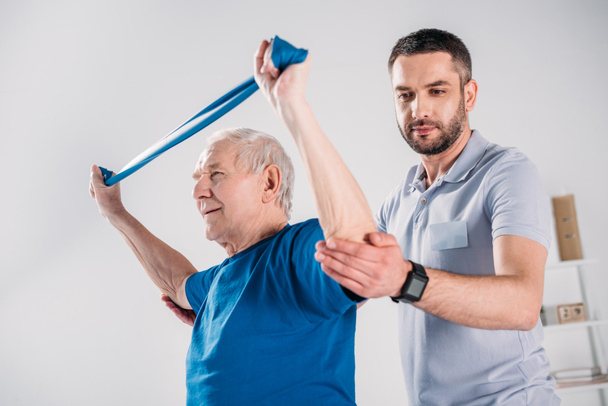 Zijaanzicht van de revalidatie-therapeut helpen senior man uitoefenend met rubber band - Foto, afbeelding