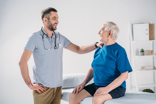 rehabilitation therapist with stethoscope and smiling senior man on massage table - Foto, afbeelding