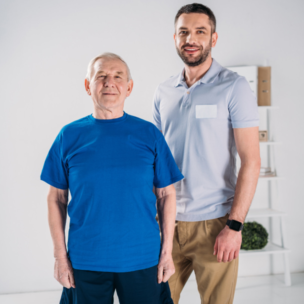 portrait of smiling senior man and rehabilitation therapist looking at camera on grey backdrop - Foto, Imagen