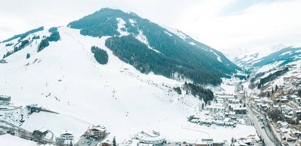 Aerial view of the mountain ski resort village in Austrian Alps with large mountain slopes, skiers, snowboarders, ski lifts and small winter town. - Φωτογραφία, εικόνα