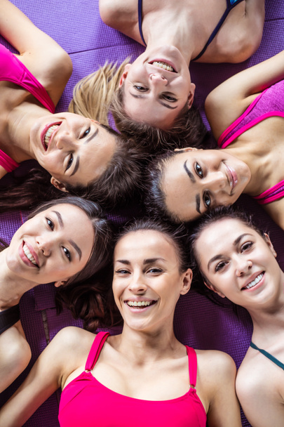 Sports friendship. Six beautiful smiling sportive women lie head to head on the floor in the gym and have fun looking at the camera - Photo, Image