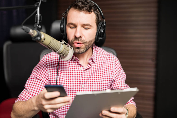 молодий латинський чоловік сидить біля мікрофона, тримає буфер обміну і використовує смартфон на радіостанції
 - Фото, зображення