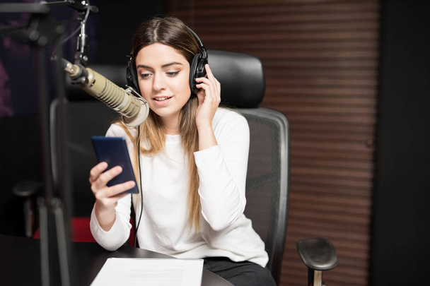 Giovane ospite femminile ispanica che comunica al microfono in studio radio con cuffie e smartphone
 - Foto, immagini