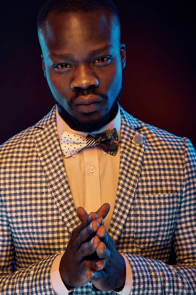 Neon portrait of handsome black man in white shirt and plaid jacket with floral bow tie is holding hands together like praying. Orange and blue studio light - Fotografie, Obrázek