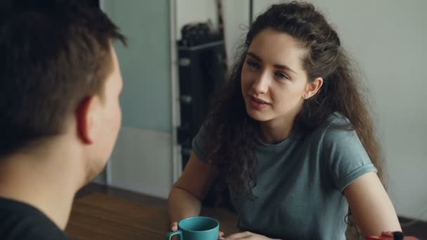 young caucasian couple sitting at table in modern kitchen dicussing something in positive way, man is sitting back to camera they are holding cups drinking tea - Footage, Video