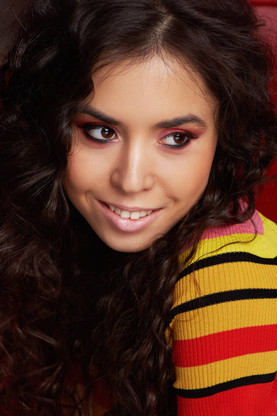 Studio fashion shooting. The girl in a bright outfit, top and pants flared in red and orange colors. Brunette with curly hair. Close-up portrait. Brown eye - Foto, afbeelding
